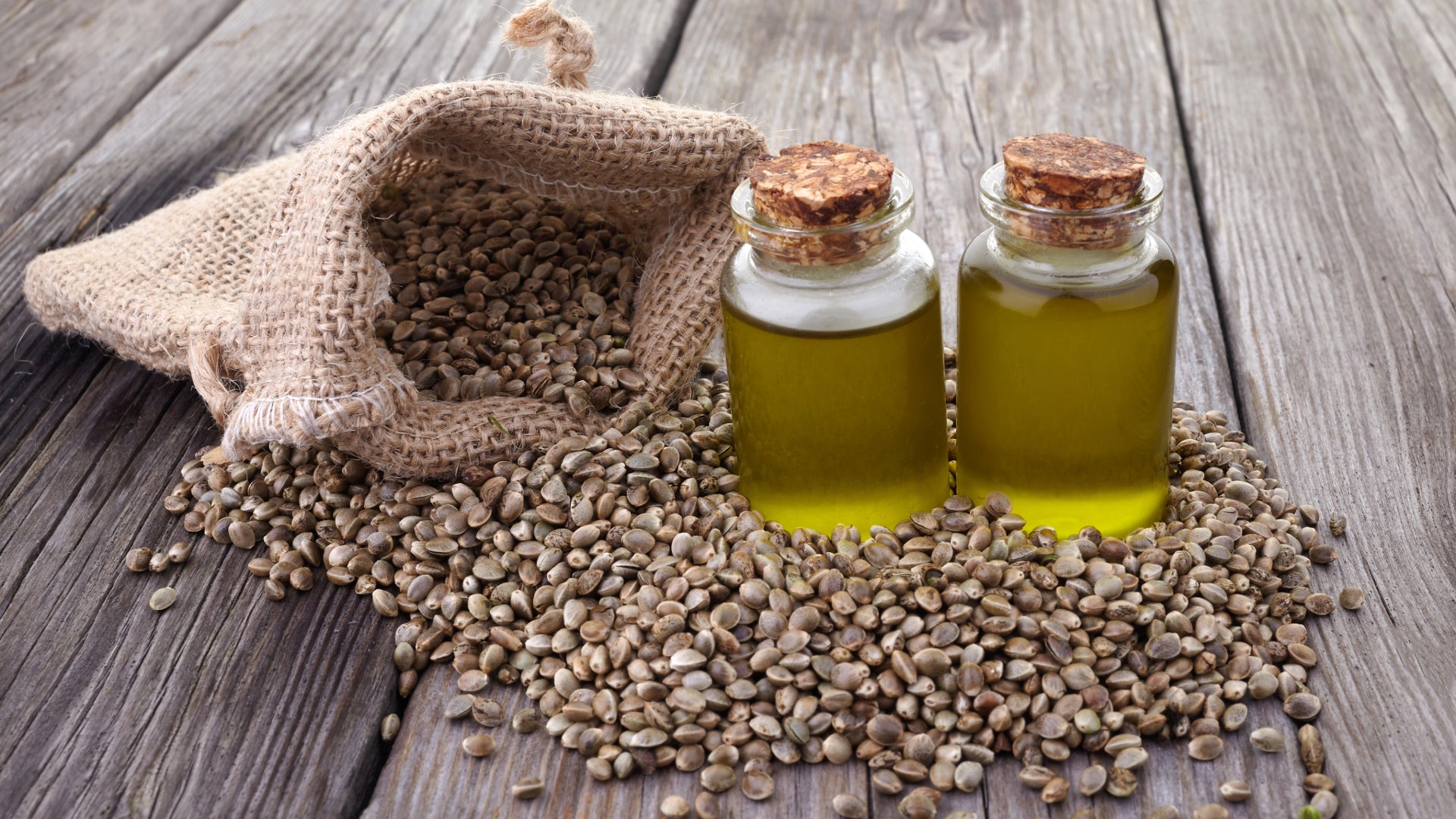 A hessian bag of hemp seeds, a wooden bowl of crushed hemp seeds and jar of hempseed oil.